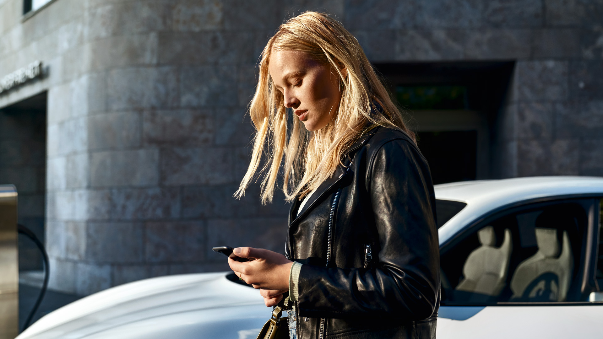 A young girl is standing in front of a Porsche with her cell phone in her hand.